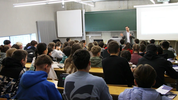 A packed lecture hall full of students from the back, a lecturer in a gray suit stands at the front and speaks to the participants.