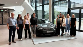 A group of people stand around a dark gray Audi RS e-tron GT, with the stairwell and windows of a modern company building in the background