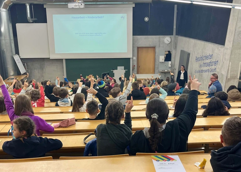 Man sieht einen mit Kindern vollbesetzten Hörsaal von hinten, einige Kinder melden sich, die Professorin schaut ins Publikum.