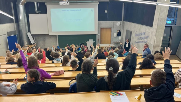 Man sieht einen mit Kindern vollbesetzten Hörsaal von hinten, einige Kinder melden sich, die Professorin schaut ins Publikum.