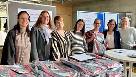  Seven ladies smile into the camera, the backpacks for the first semester students lined up in front of them. 