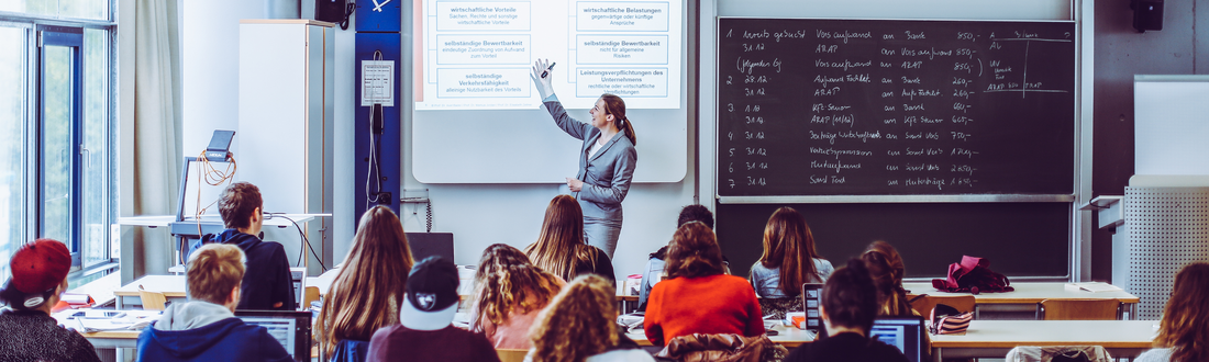 Eine Dozentin spricht vor Studenten und zeigt auf Inhalte, die ein Overheadprojektor auf die Wand wirft 