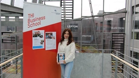 A woman next to a movable display on which two posters are hanging, holding a tablet showing an Instagram post, the THI's glass façade in the background.